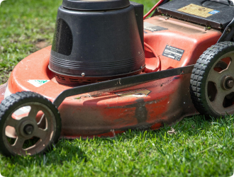 Close-up of a red lawn mower cutting lush green grass. The mower's worn, curved body and black wheels suggest frequent use, conveying a sense of routine maintenance.