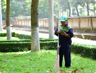 A person in a green cap and uniform stands on lush grass in a park, holding a broom and looking at a phone. Trees and a path are in the background.