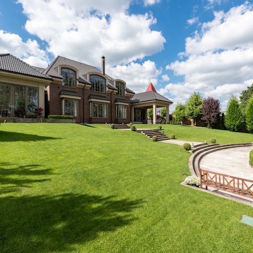 Expansive brick house with large windows, surrounded by vibrant green lawn and trees under a bright blue sky with fluffy white clouds.