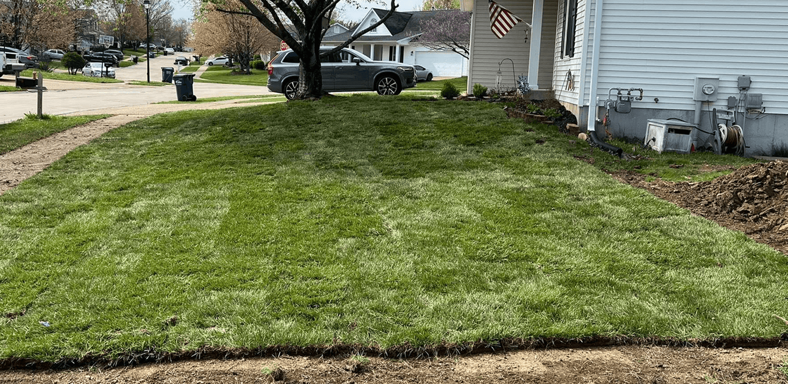 Freshly laid green sod covers a front yard, bordered by a white house. A tree stands on the left, with a street and parked cars in the background.