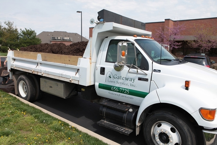 A white dump truck with "Gateway Lawn Service, Inc." branding is parked on a road. The truck bed is filled with dark soil. Nearby are blooming trees and a brick building.