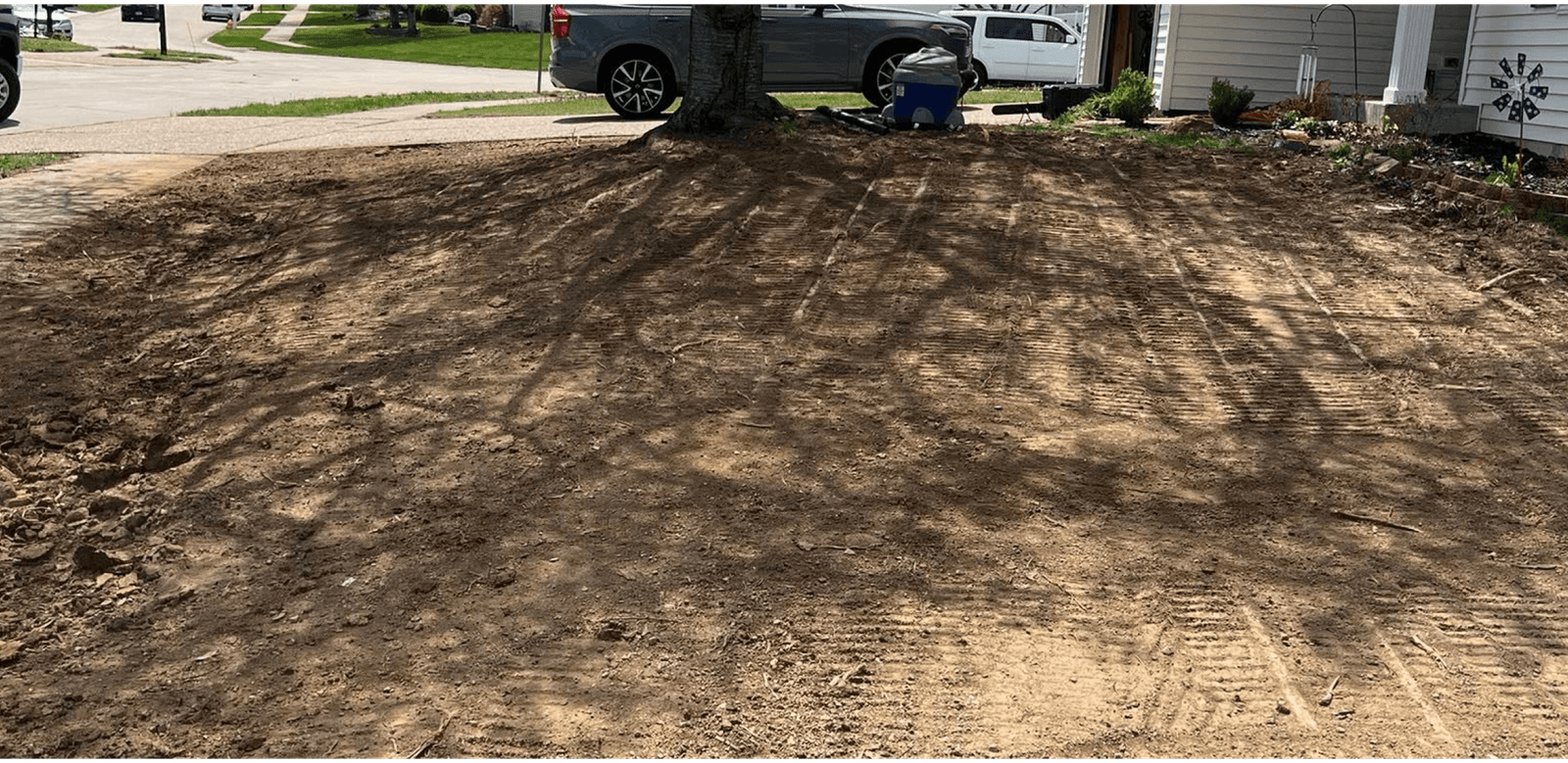 Freshly tilled earth in a front yard casts shadows from nearby trees, with a parked car and residential houses visible in the background.