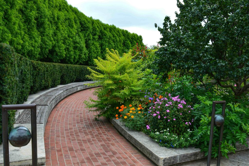 A serene garden path with a curved brick walkway, lush greenery, and colorful flowers. Hedgerow in the background, creating a peaceful ambiance.