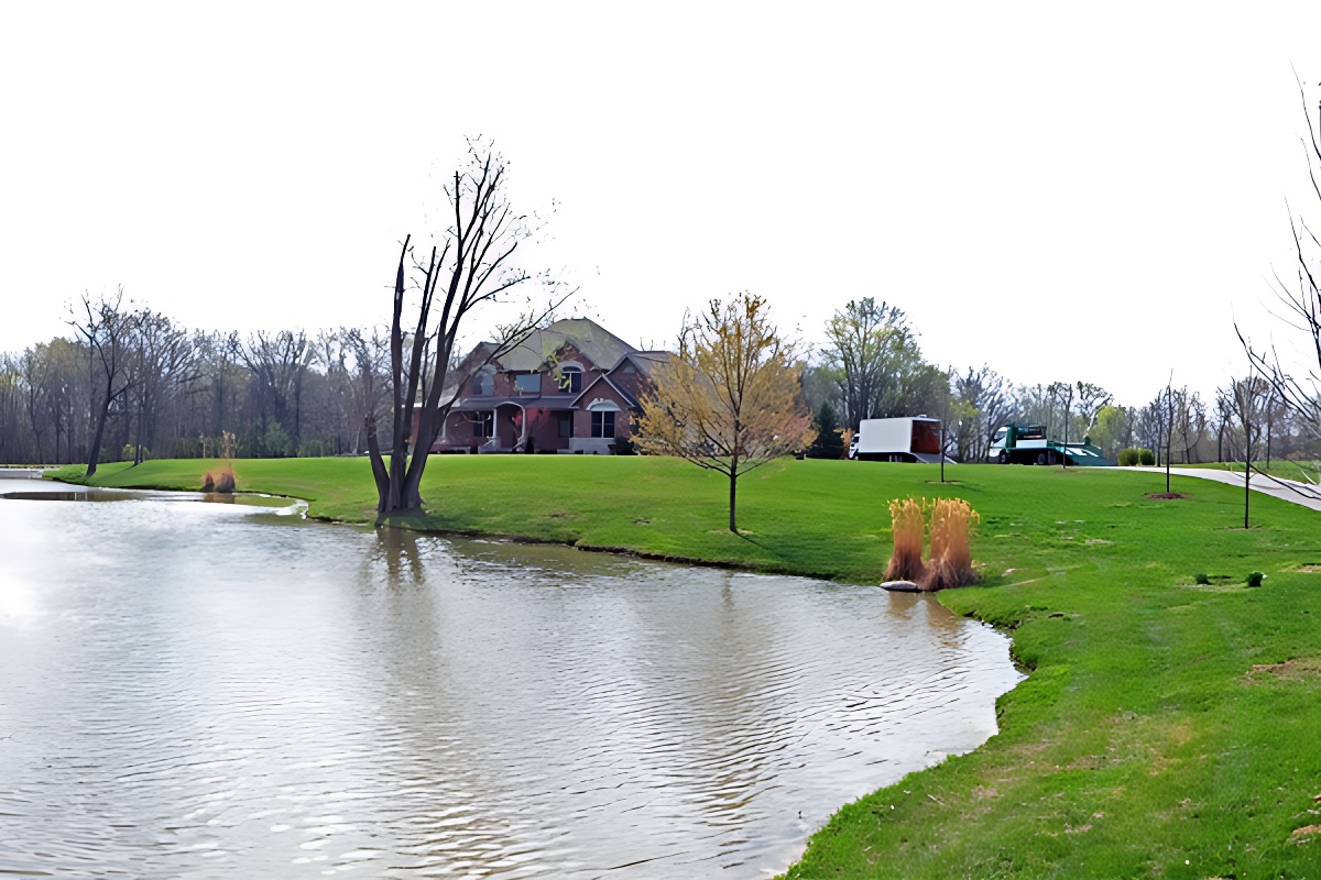 A serene landscape with a large house surrounded by lush green grass and trees. A tranquil pond reflects the overcast sky in the foreground.