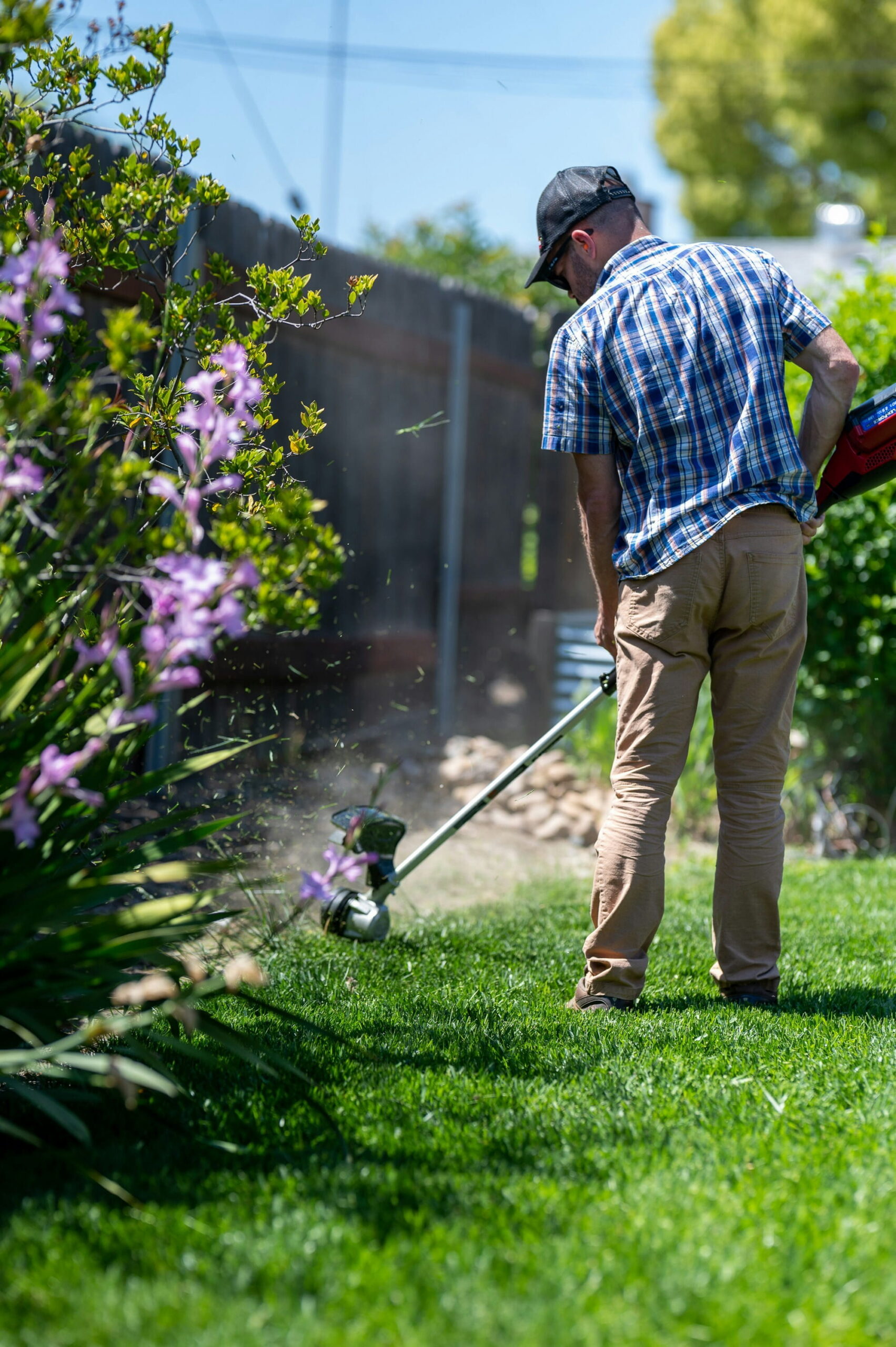 Man in a blue plaid shirt and cap trims grass with a string trimmer in a sunny garden. Vibrant flowers and lush greenery surround him.