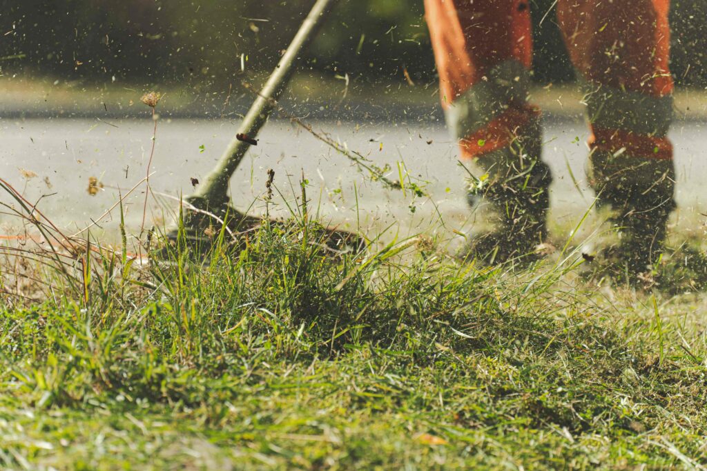 A worker in orange safety gear uses a weed trimmer on tall grass, with clippings flying in the air, suggesting motion and action on a sunny day.