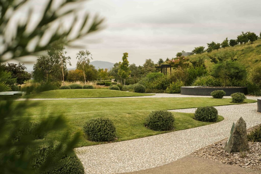 Manicured garden with lush green grass, circular shrubs, and a winding pebble path. Trees and a distant hill create a serene, peaceful setting.
