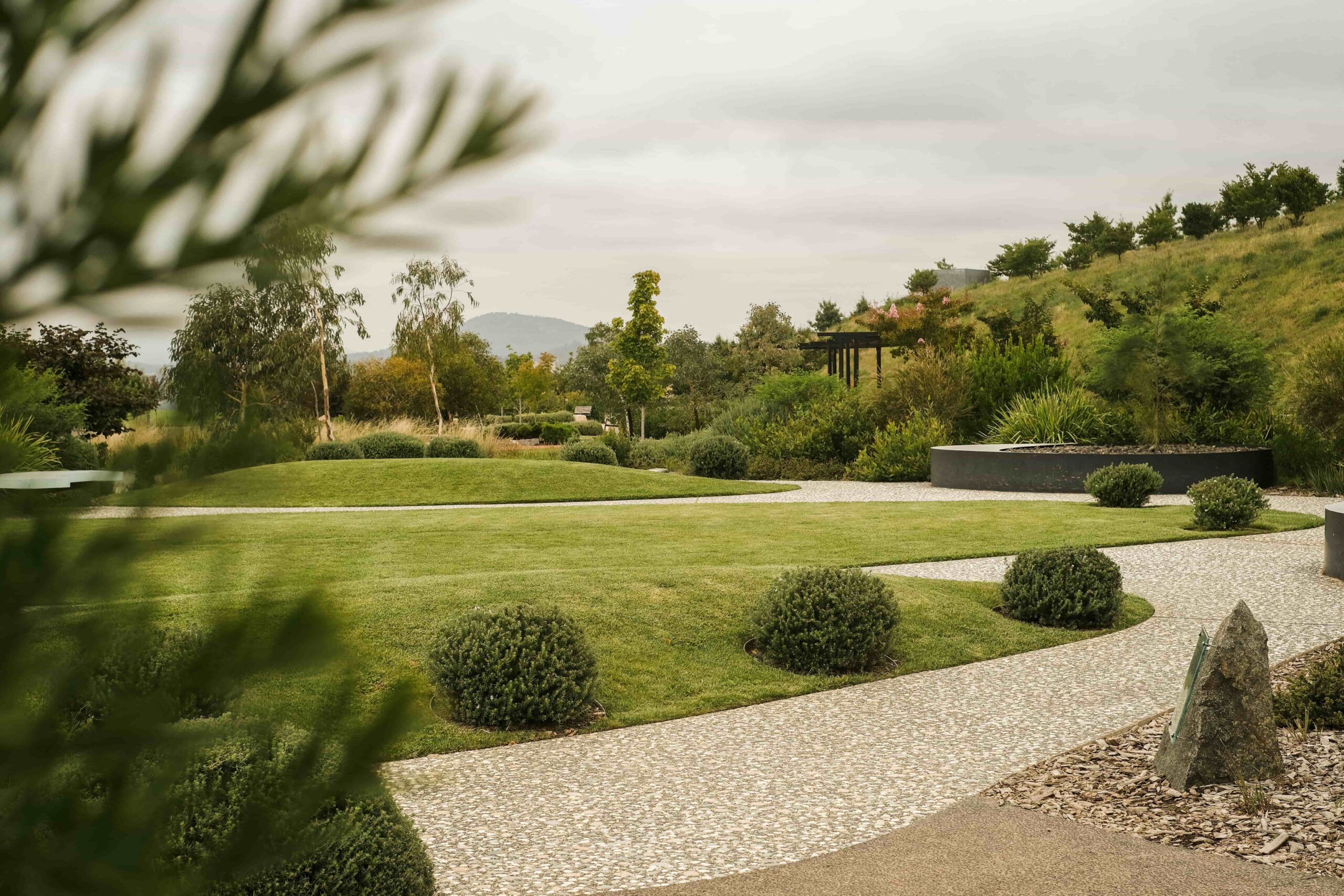 Manicured garden with lush green grass, circular shrubs, and a winding pebble path. Trees and a distant hill create a serene, peaceful setting.