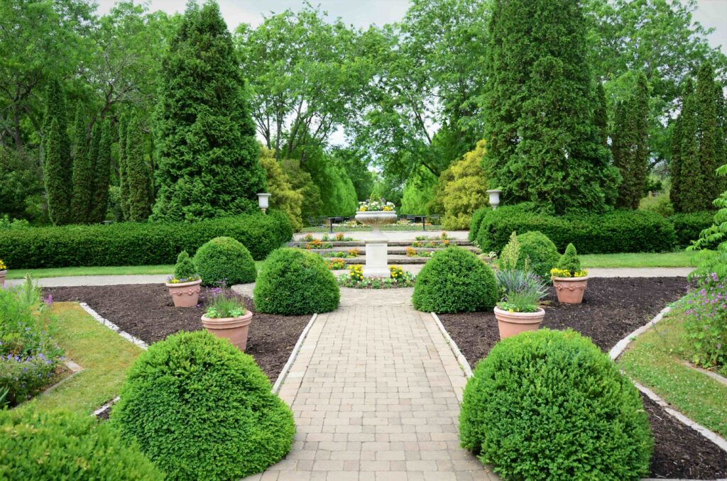 Elegant formal garden with a stone pathway, neatly trimmed hedges, potted plants, and a central fountain. Lush greenery creates a serene ambiance.