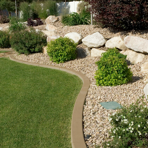 A well-manicured garden featuring a curved border separating lush green grass from a gravel area with scattered bushes and large rocks.